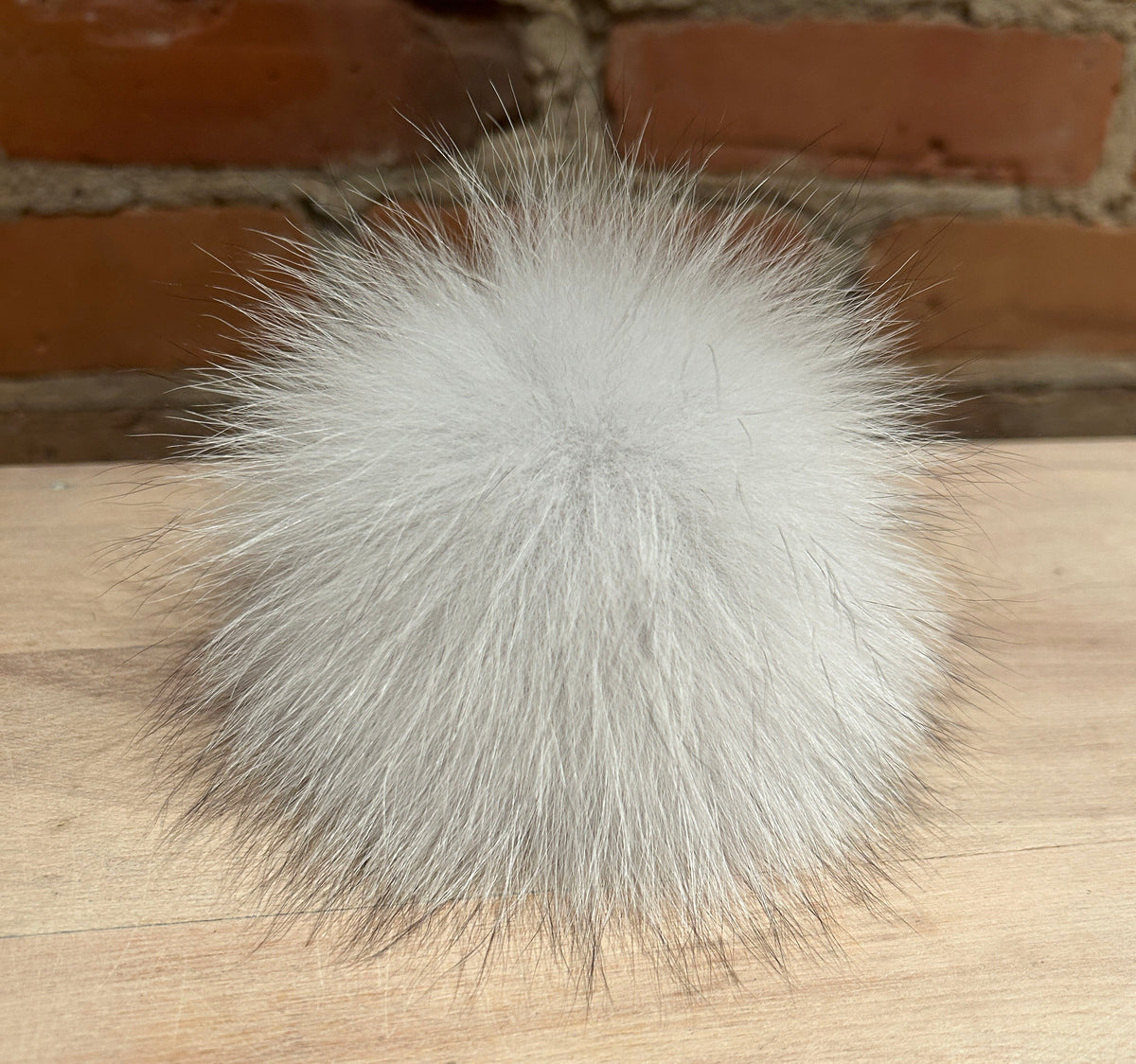 A fluffy white fox fur pom-pom ball resting on a light wood surface with a brick wall background.