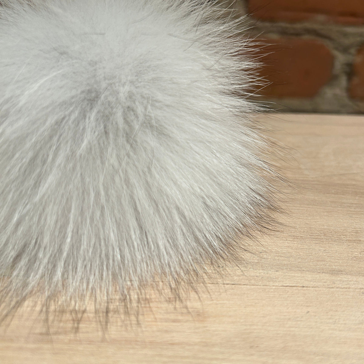 A close-up of a fluffy white fox fur pom-pom resting on a light wood surface with a blurred brick wall background.