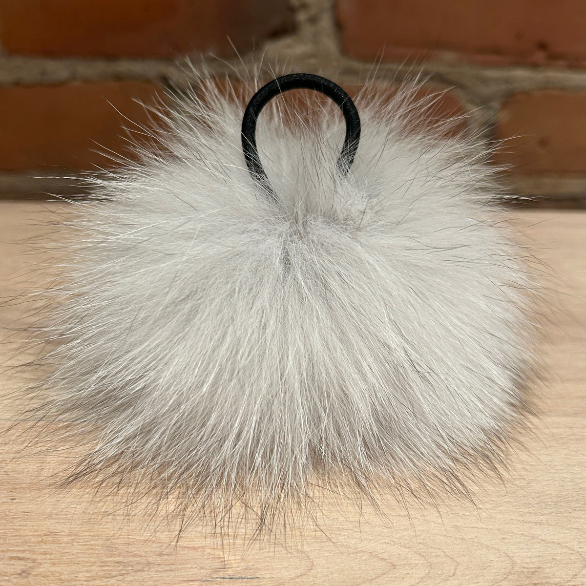 A fluffy light gray fox fur pom-pom bag charm with a black elastic loop, resting on a light wood surface with a brick wall background.