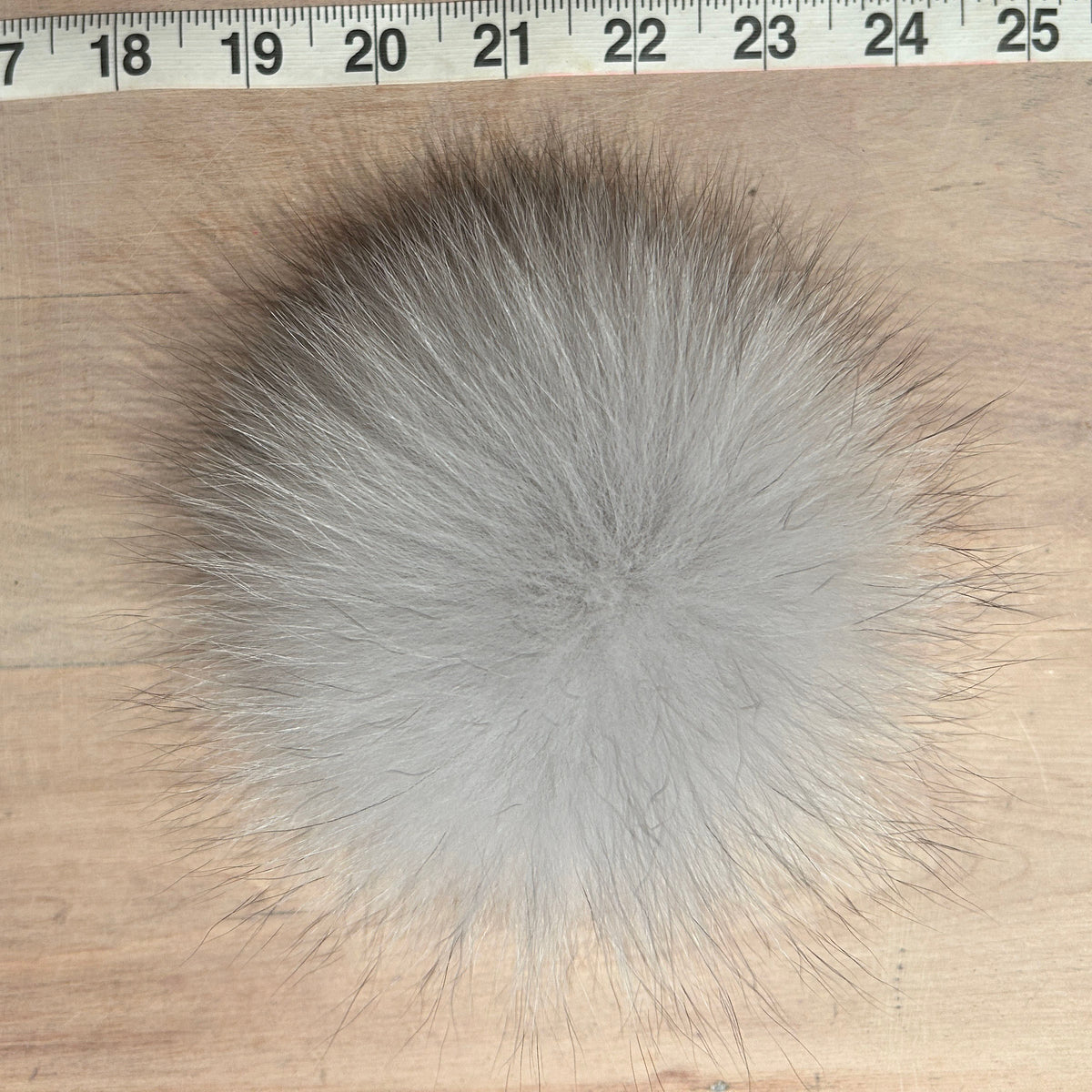 A round, fluffy fox fur pom-pom in light gray and white, shown on a light wood surface with a measuring tape ruler at the top for scale.