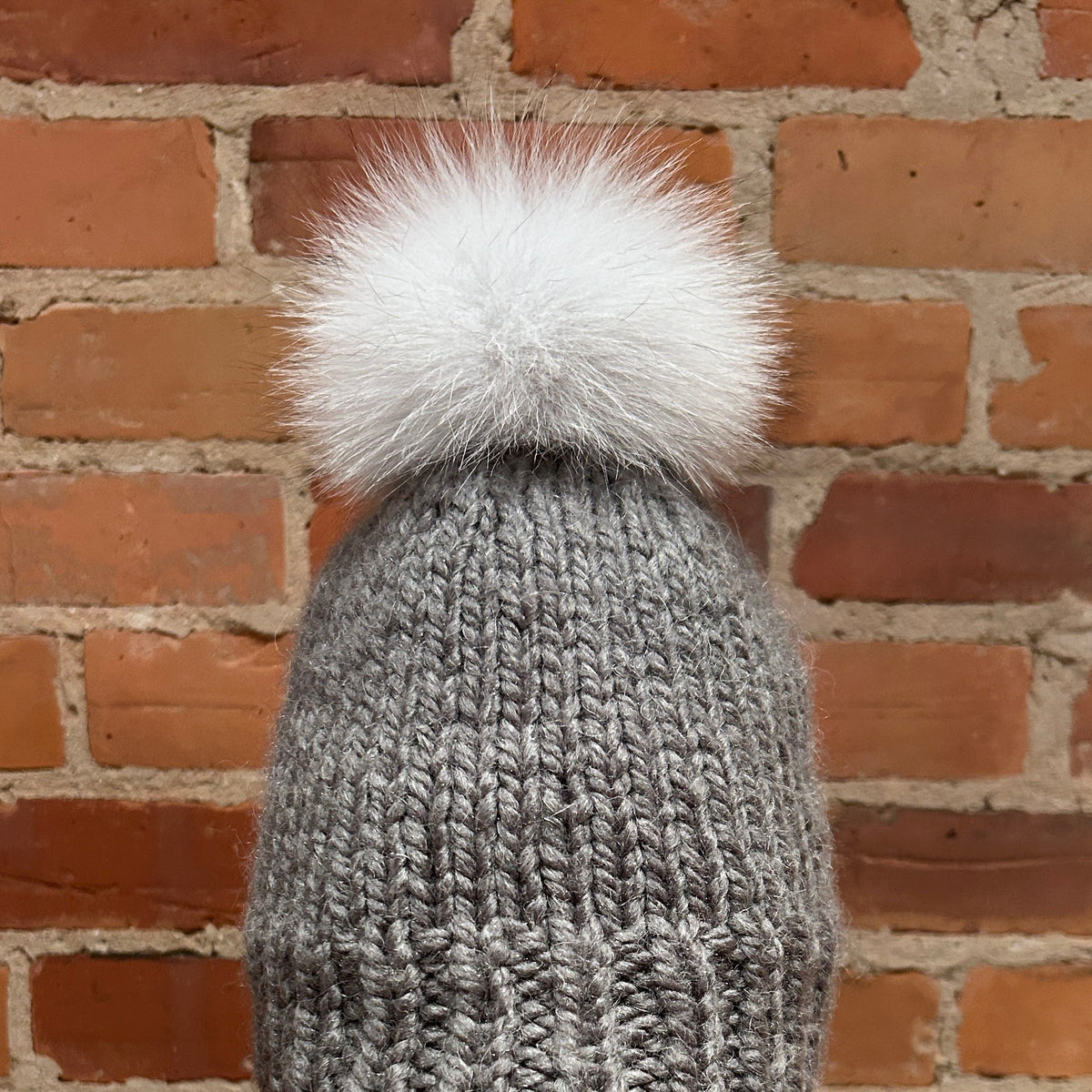 A gray chunky knit beanie with a large white fox fur pom-pom, displayed on a mannequin head with red lipstick against a red brick wall background.