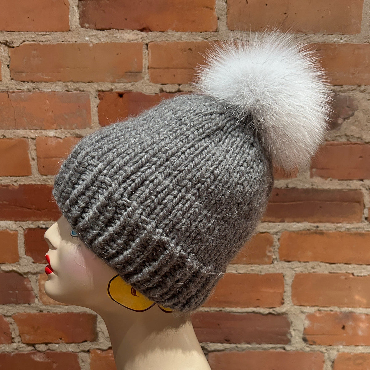 A gray ribbed knit beanie with a large fluffy white pom-pom, displayed on a mannequin head against a red brick wall background.