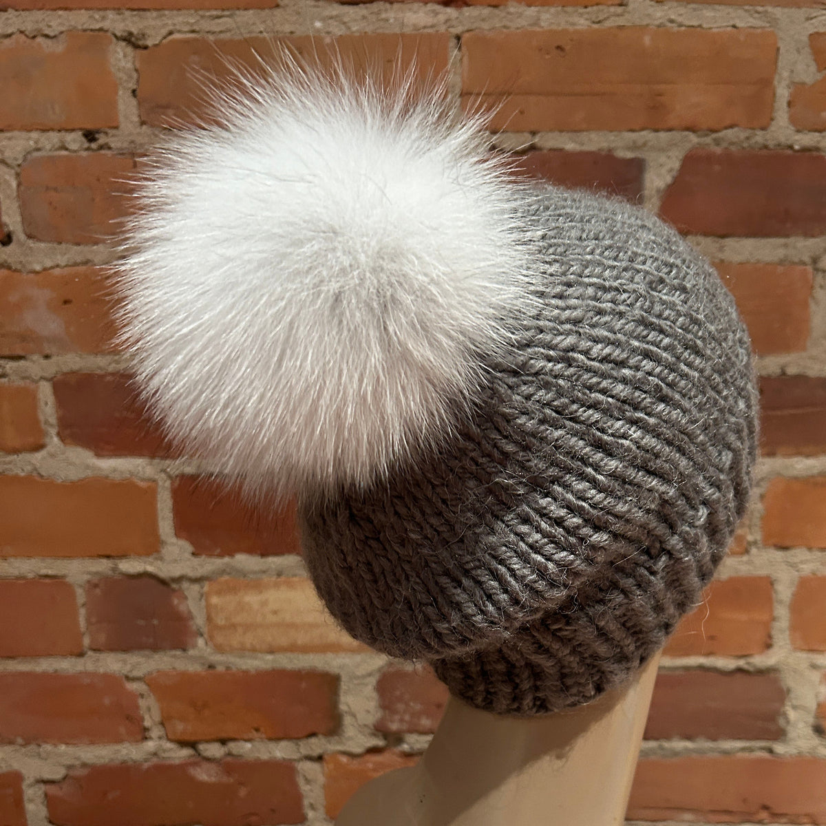 A gray ribbed knit beanie with a large fluffy white fox-fur pom-pom, displayed on a mannequin head against a red brick wall background.