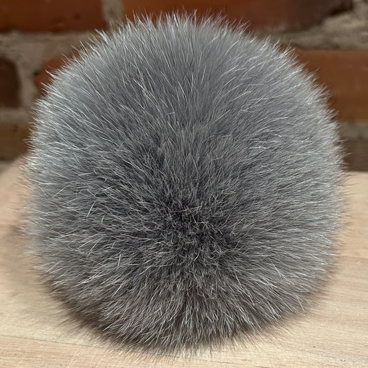 A round, fluffy gray fox-fur pom-pom ball resting on a light wood surface with a blurred brick wall in the background.