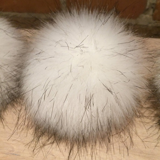 A close-up of a fluffy white faux fur pom-pom with dark guard hairs, resting on a light wood surface with a brick wall in the background.