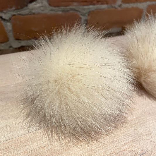 A close-up of a fluffy cream-colored fox fur pom-pom resting on a light wood surface with a brick wall in the background.