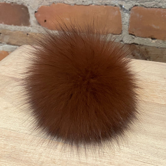 A round, fluffy brown fox fur pom-pom accessory resting on a light-colored wooden surface with a brick wall background.