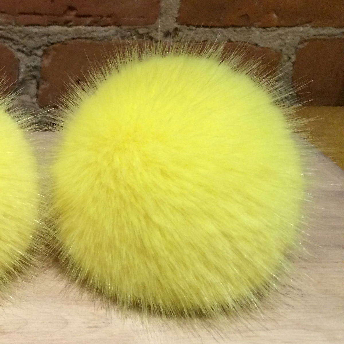 A bright yellow fluffy faux fur pom-pom ball resting on a light wood surface with a brick wall background.