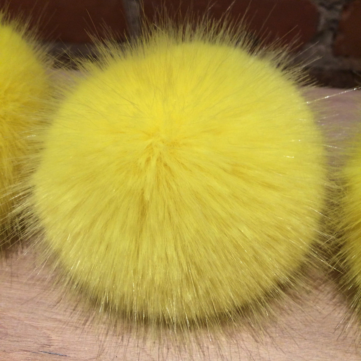 A bright yellow fluffy faux fur pom-pom ball resting on a wooden surface, with other similar yellow balls partially visible in the background.