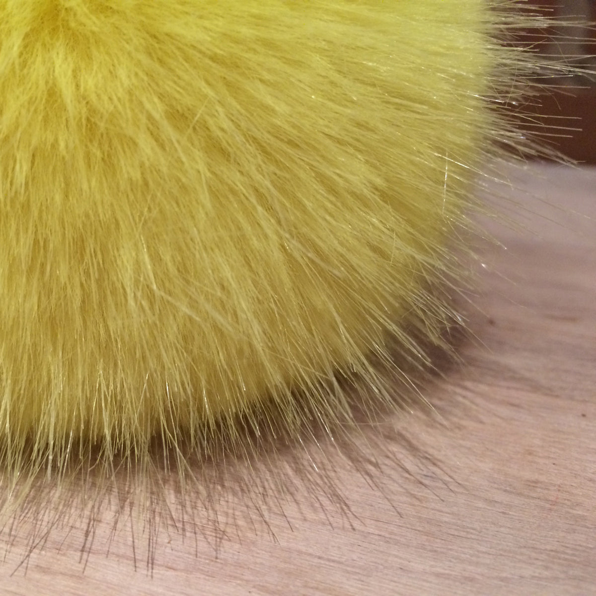 Close-up of a bright yellow faux fur pom-pom resting on a light wood surface.