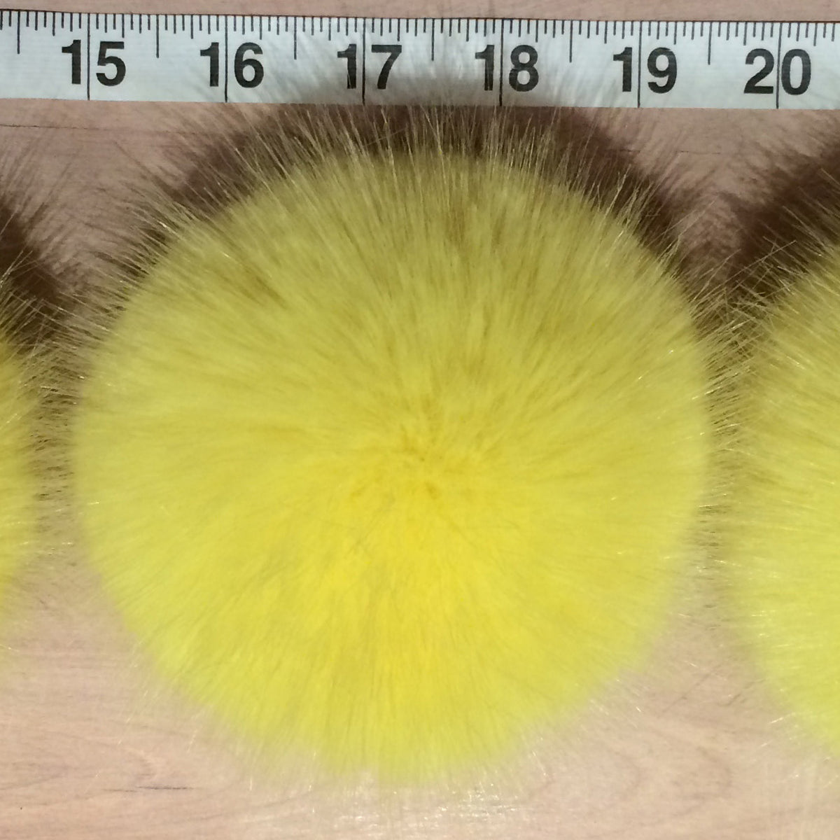 A close-up of a round, fluffy yellow faux fur pom-pom with a measuring ruler visible at the top to indicate size, set on a light wood surface.