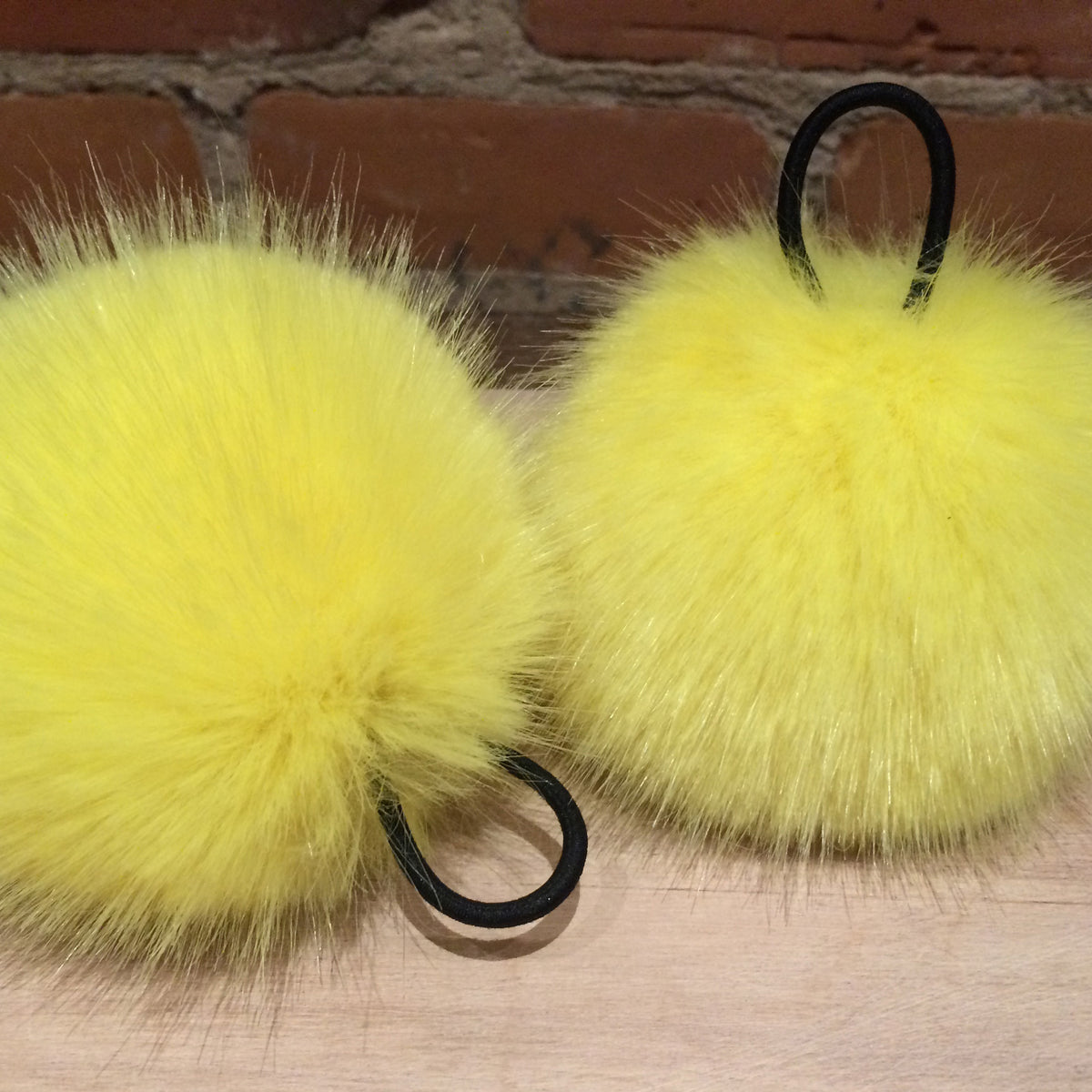 Two fluffy bright yellow faux fur pom-pom keychains with black elastic loops, resting on a light wood surface with a brick wall background.
