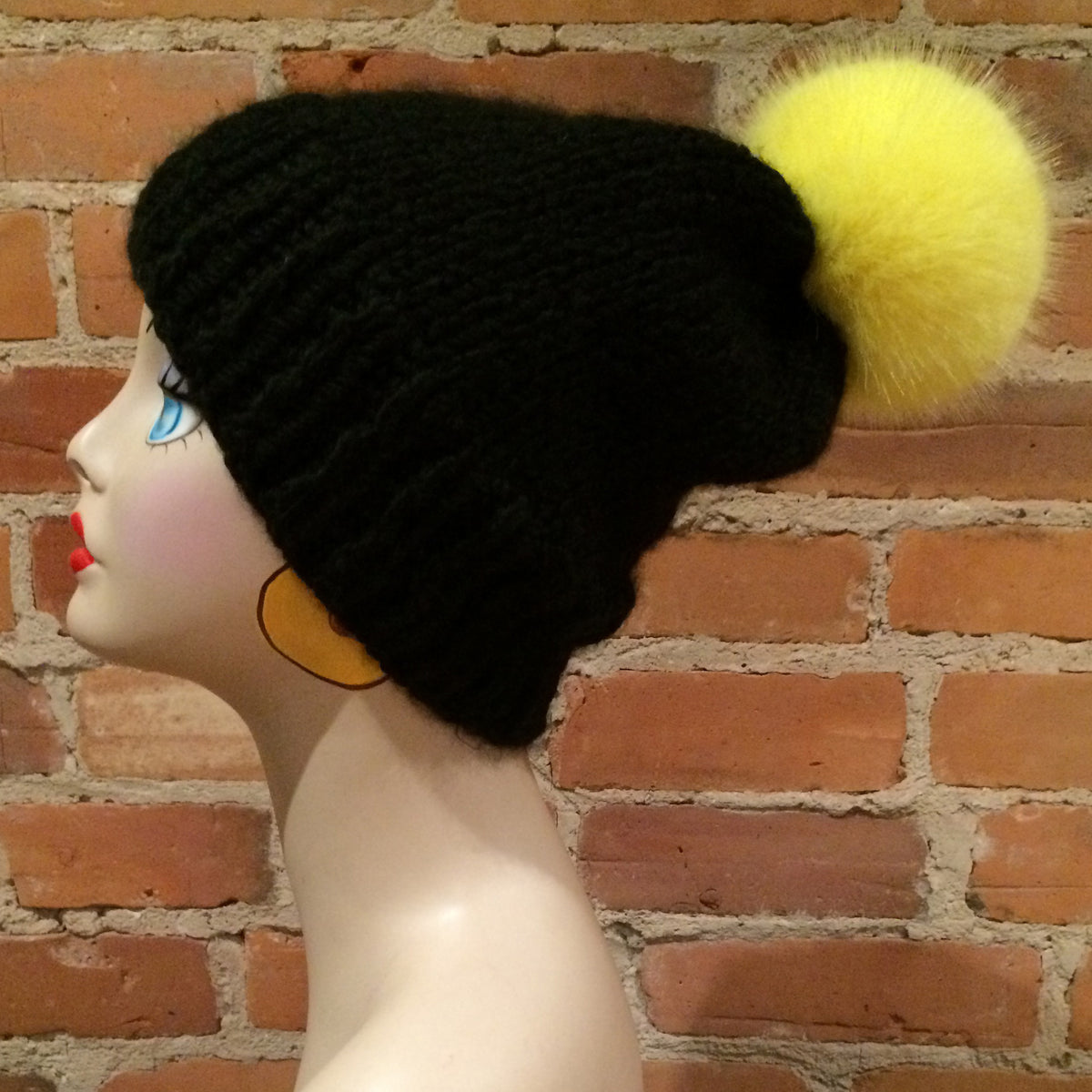 A black knit beanie hat with a fluffy yellow pom-pom, displayed on a mannequin head against a red brick wall background.