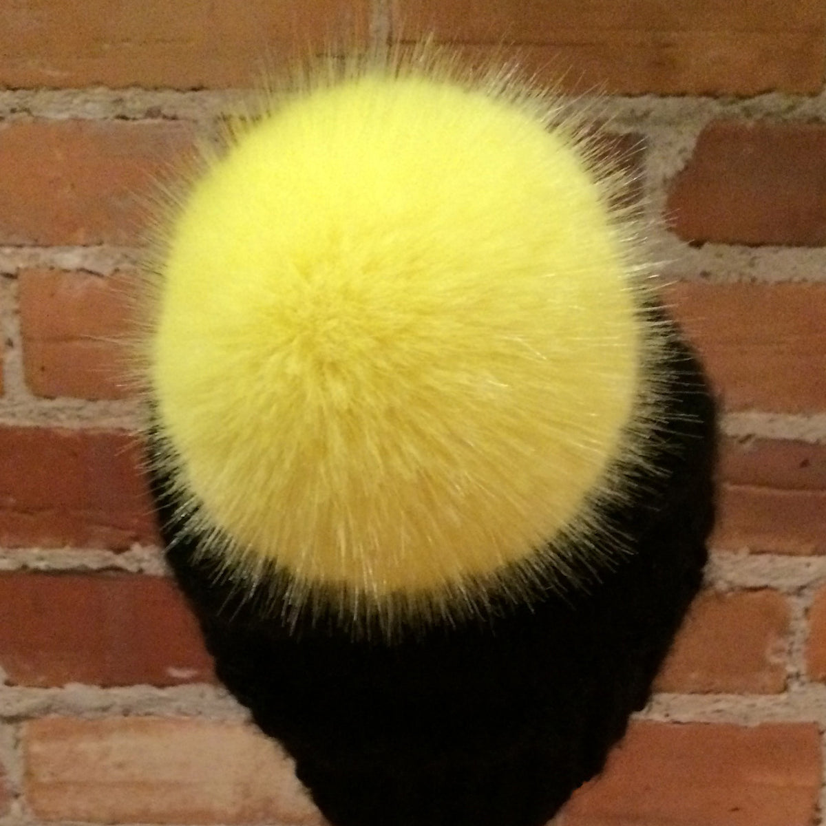 A black knit beanie hat featuring a large, fluffy yellow faux fur pom-pom, displayed against a red brick wall background.