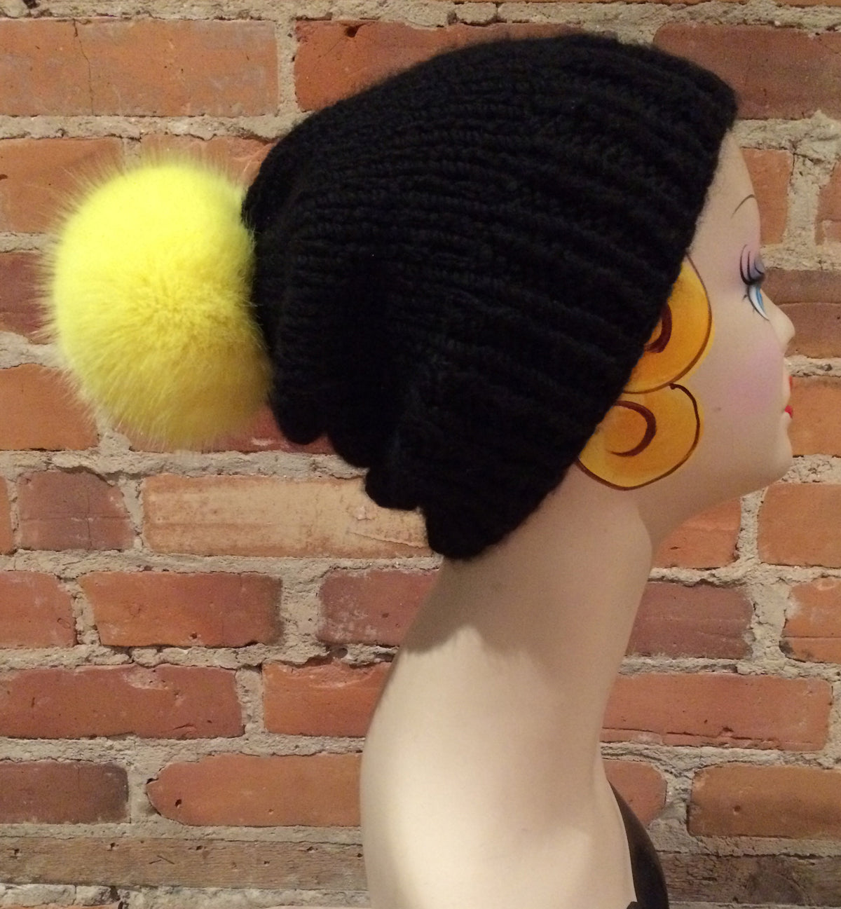 A black ribbed knit beanie hat with a fluffy yellow pom-pom, displayed on a mannequin head against a red brick wall background.