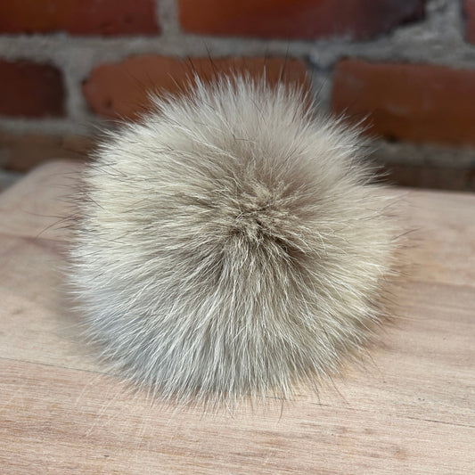 A fluffy coyote-fur pom-pom accessory in a light beige and gray color, resting on a light wood surface with a brick wall background.