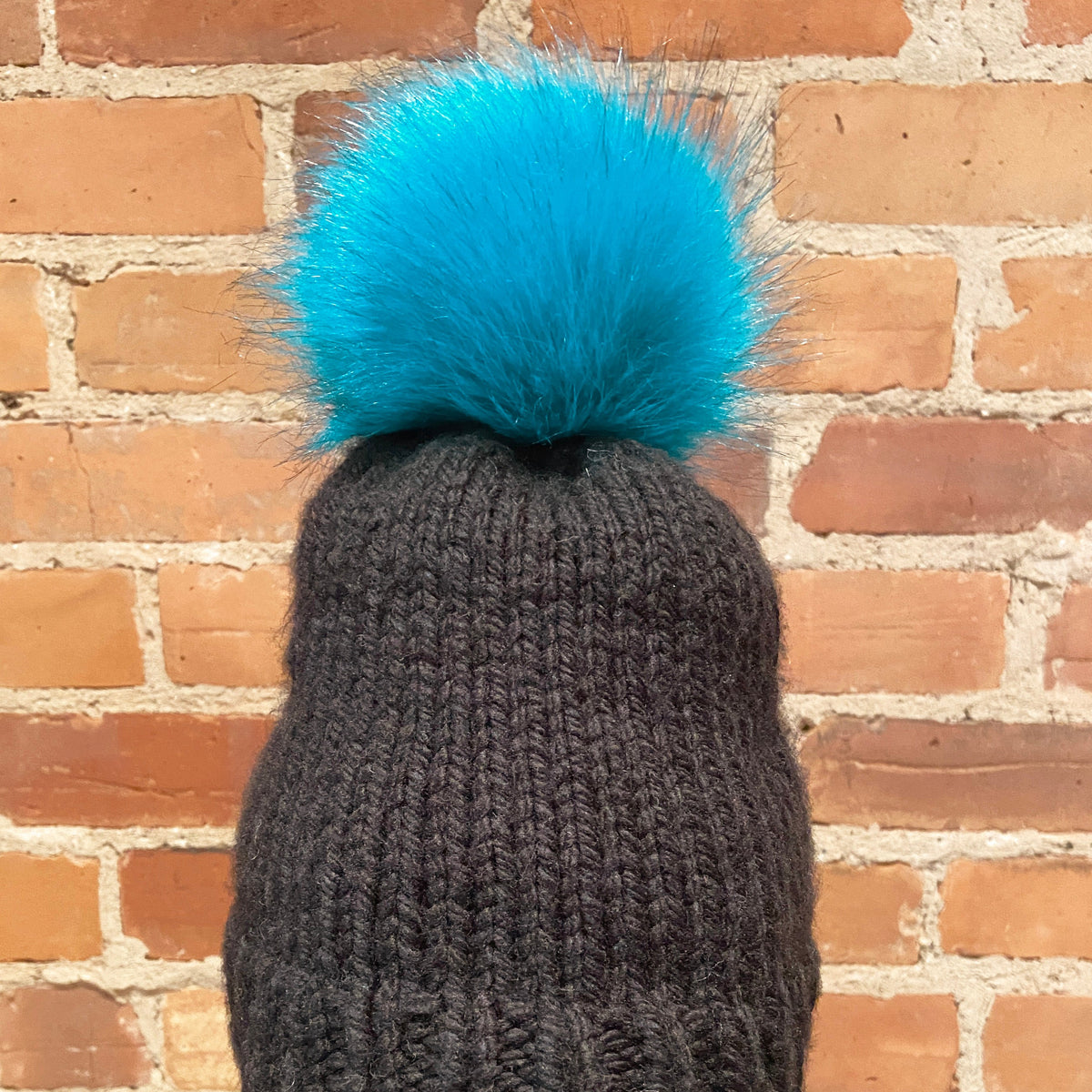 A dark charcoal gray ribbed knit beanie with a large, fluffy bright turquoise blue pom-pom, displayed on a mannequin head with red lips against a brick wall background.