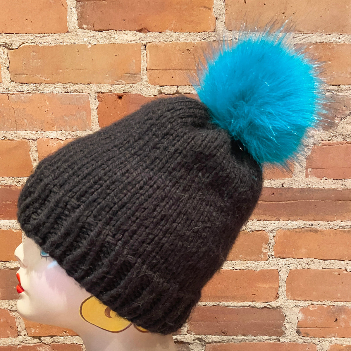 A dark charcoal gray ribbed knit beanie with a fluffy bright turquoise blue pom-pom, displayed on a mannequin head against a red brick wall background.