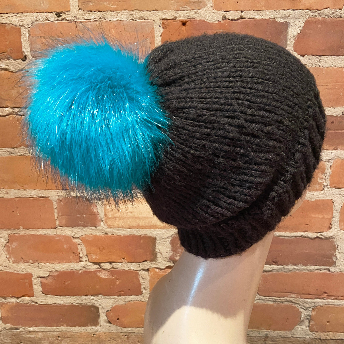 A black ribbed knit beanie with a large, fluffy bright turquoise blue faux fur pom-pom, displayed on a mannequin head against a red brick wall background.