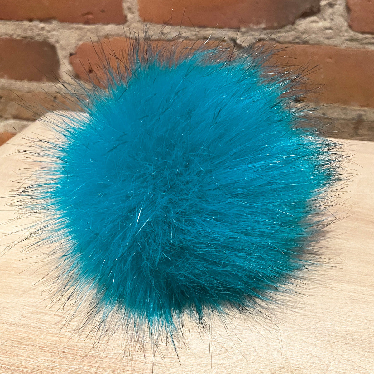 A bright turquoise blue fluffy faux fur pom-pom resting on a light wood surface with a red brick wall in the background.