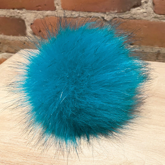 A bright turquoise blue fluffy faux fur pom-pom resting on a light wood surface with a red brick wall in the background.