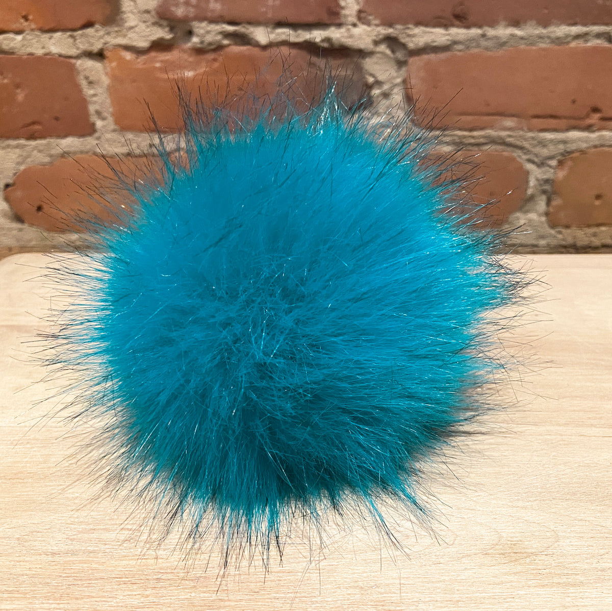 A bright turquoise blue faux fur pom-pom ball resting on a light wood surface with a red brick wall in the background.