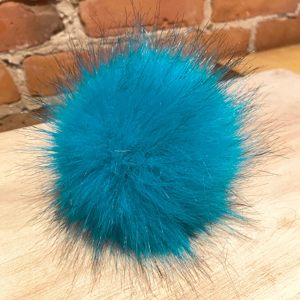 A bright turquoise blue fluffy faux fur pom-pom with dark bristles, resting on a light wood surface with a brick wall background.