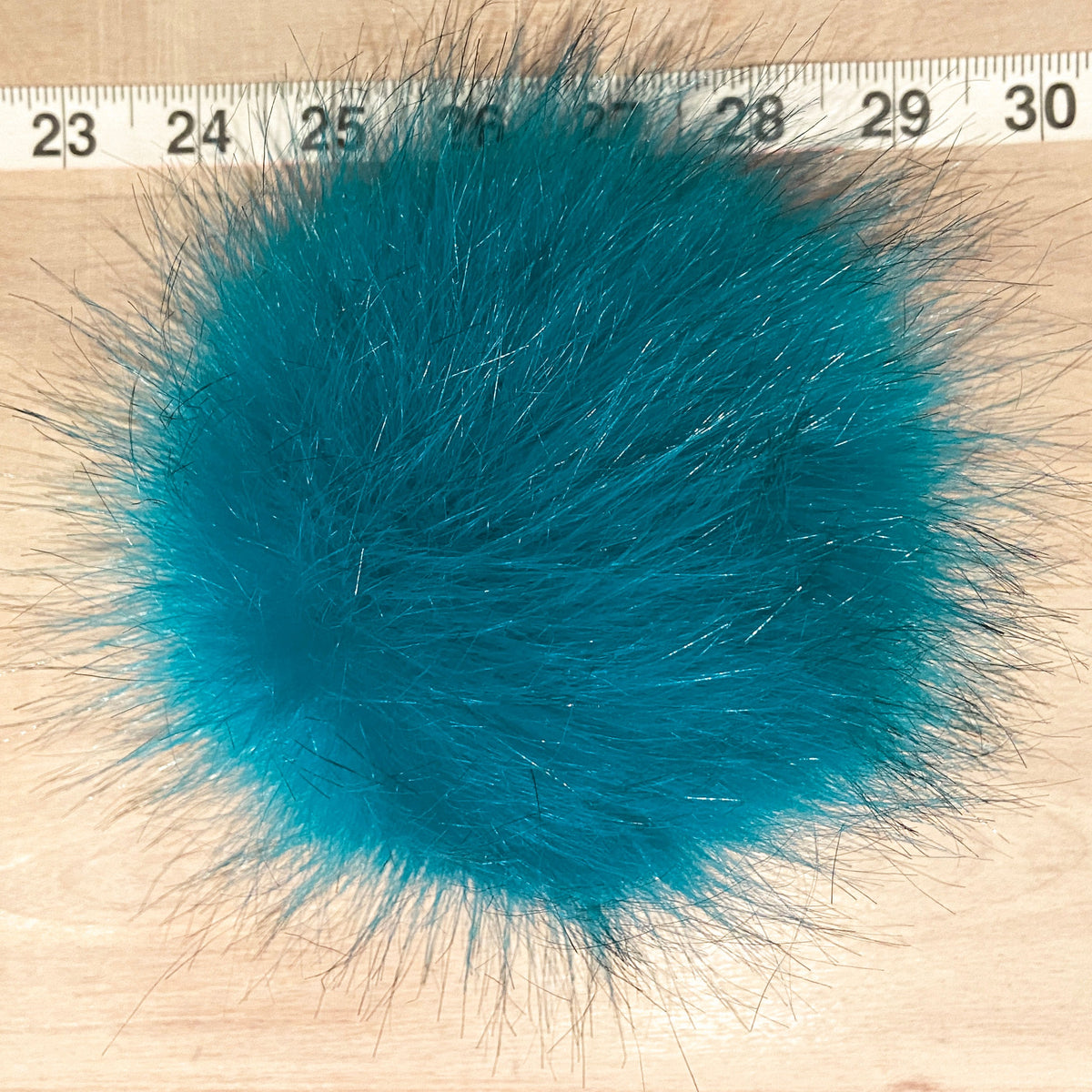 A round, fluffy faux fur pom-pom in a vibrant teal blue color, shown on a light wood surface with a measuring tape ruler at the top for scale.