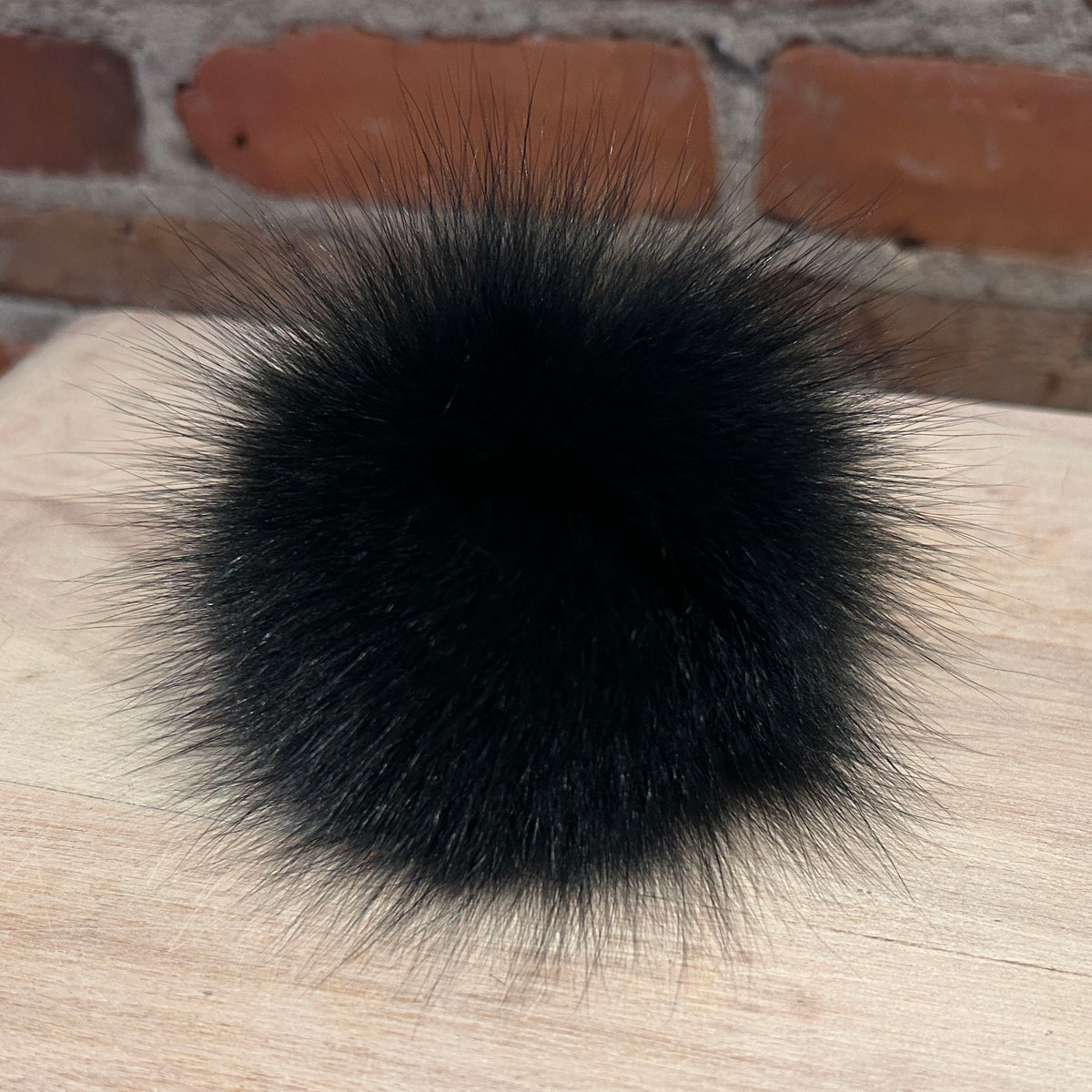 A round black fox fur pom-pom accessory resting on a light wood surface with a brick wall background.
