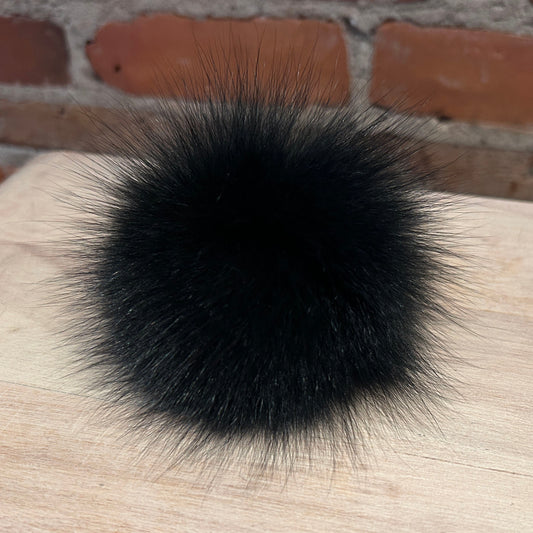 A round black fox fur pom-pom accessory resting on a light wood surface with a brick wall background.