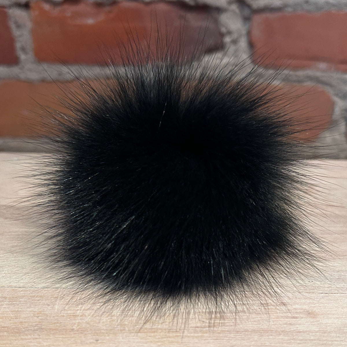 A round, fluffy black fox fur pom-pom accessory resting on a light wood surface with a blurred red brick wall background.