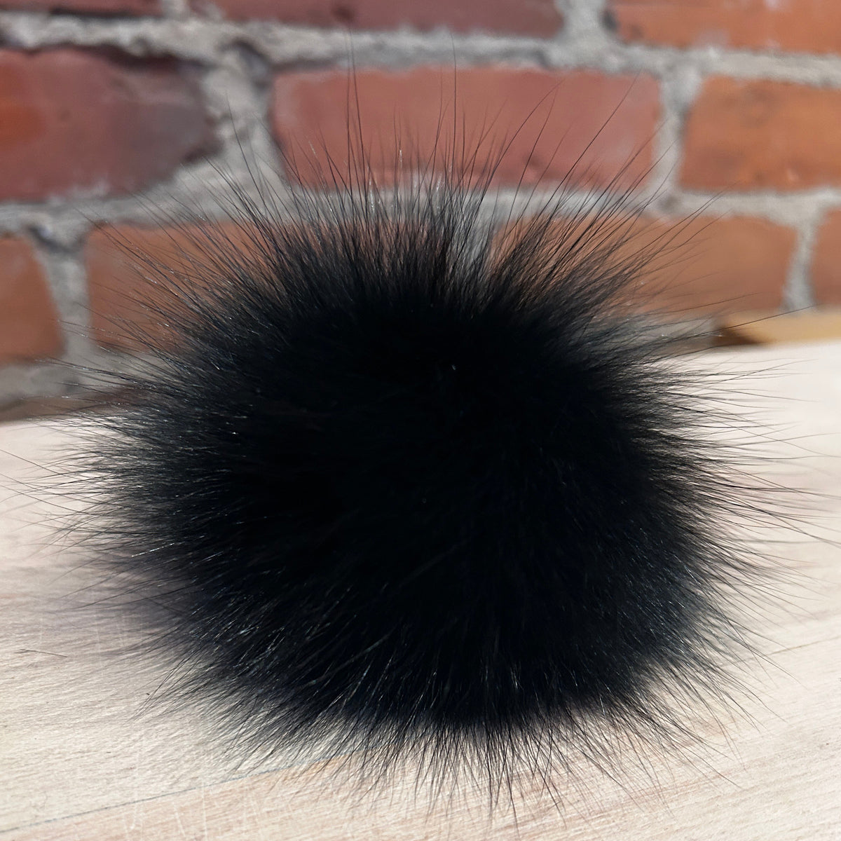A round black fur pom-pom accessory resting on a light-colored wooden surface with a blurred red brick wall background.