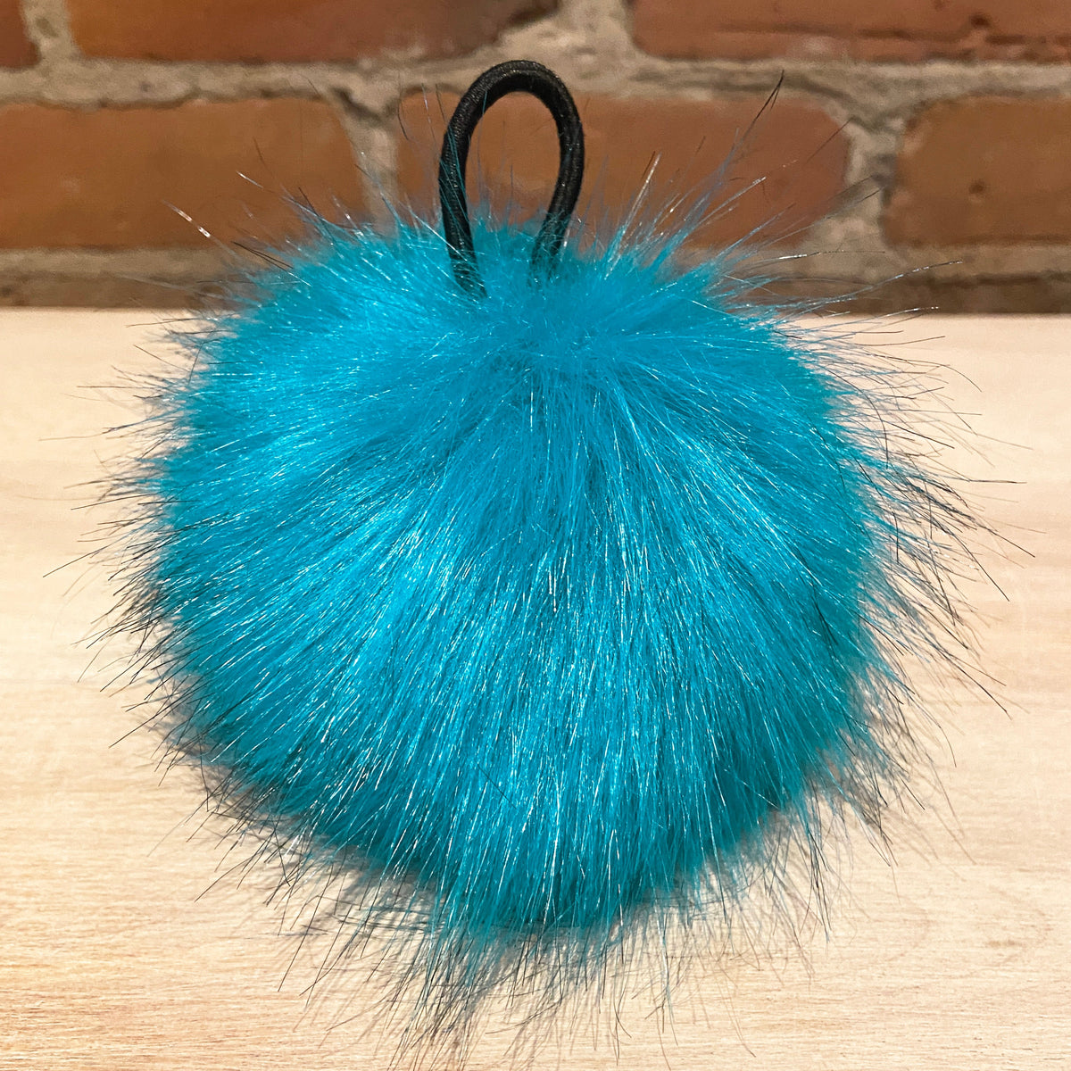 A bright turquoise blue faux fur pom-pom keychain with a black elastic loop, resting on a light wood surface with a brick wall background.