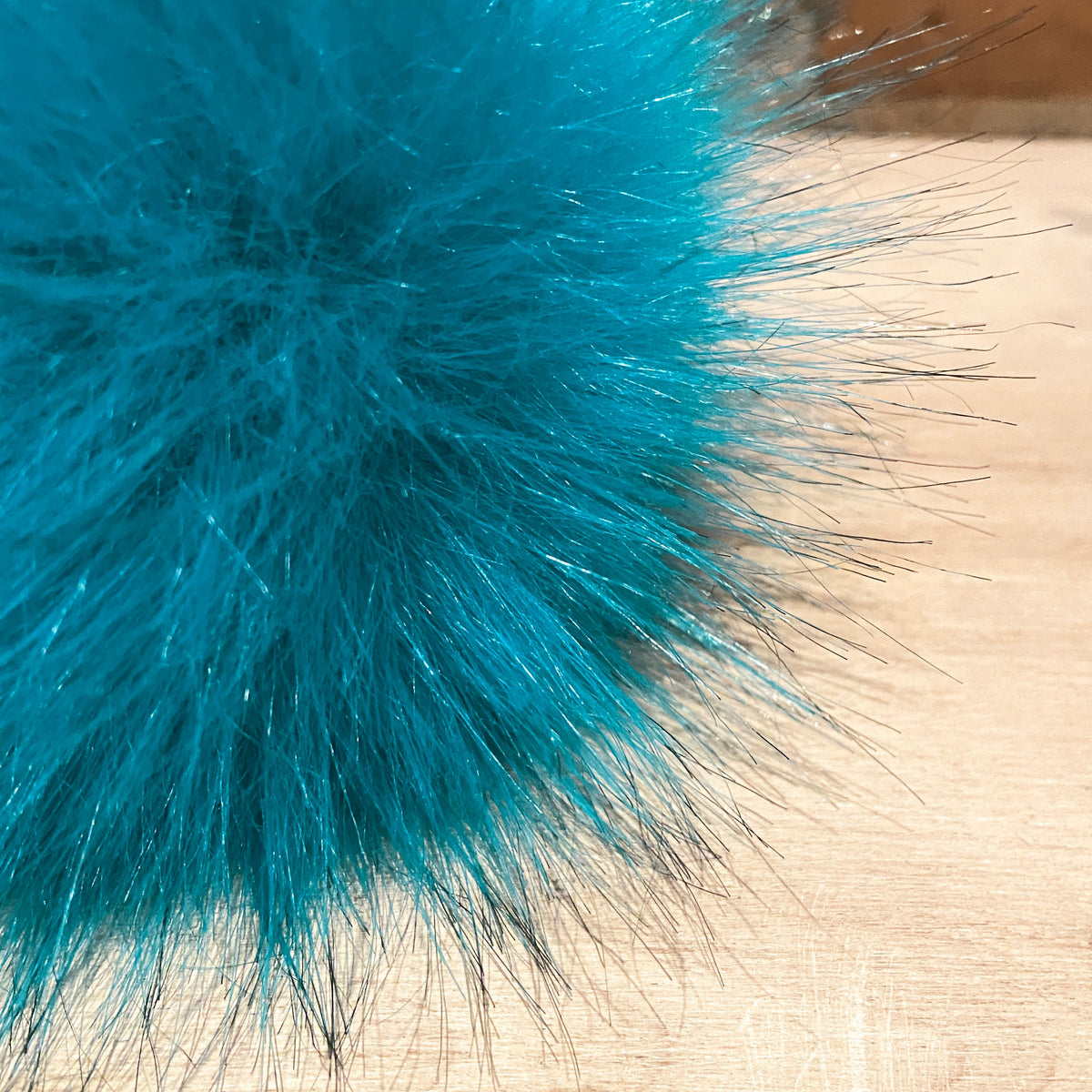 A close-up of a fluffy, electric turquoise blue faux fur pom-pom with subtle silver glitter strands, resting on a light wood surface.