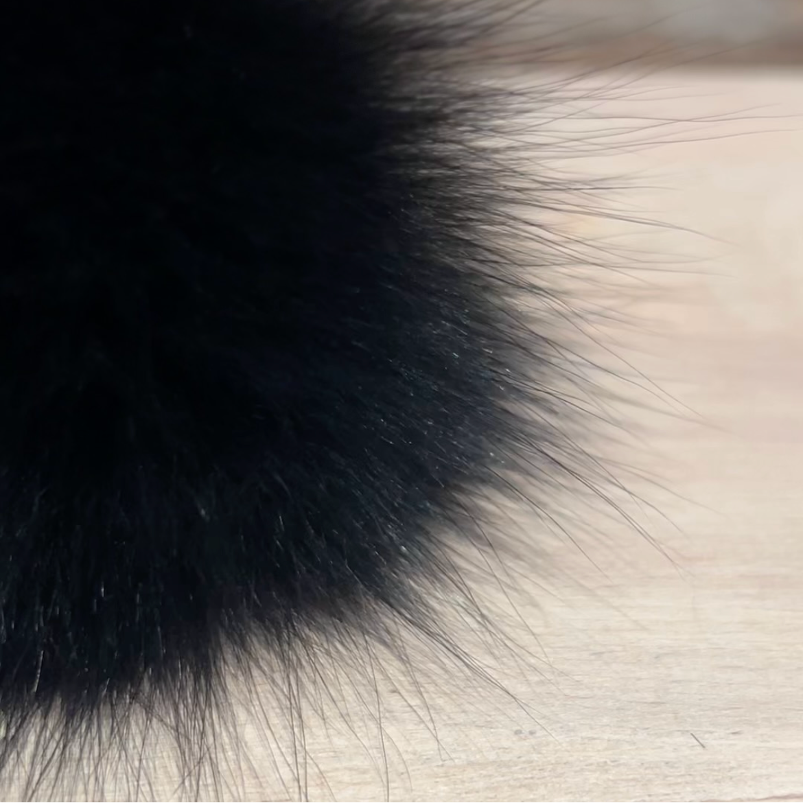 A close-up of a fluffy black fox fur pom-pom with individual strands visible against a light wood surface and blurred background.