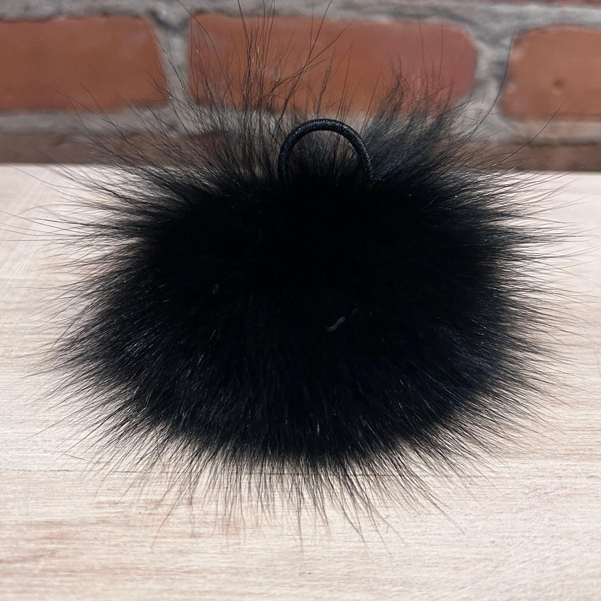 A black fox fur hat pom with black loop attachment, resting on a light wood surface with a brick wall background.