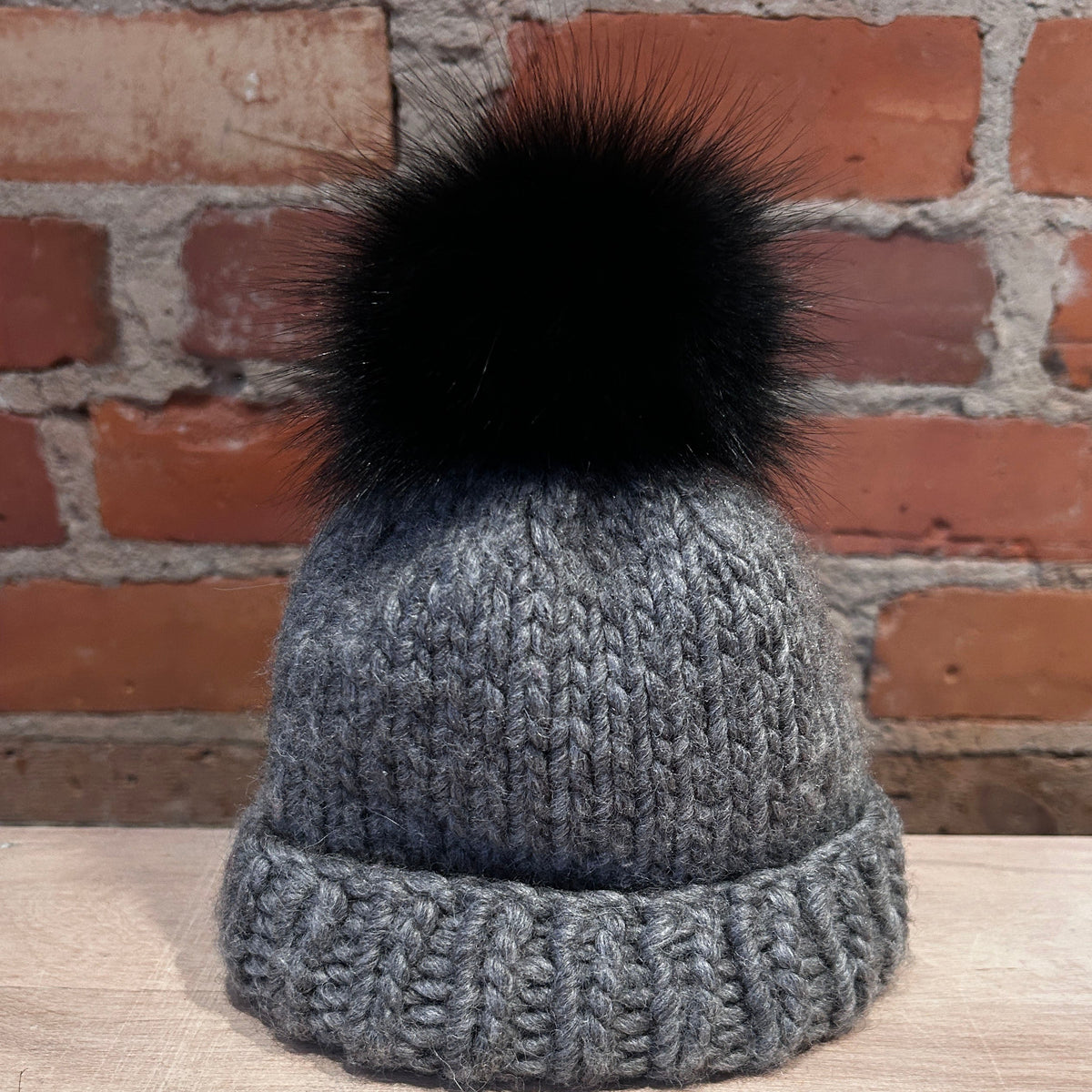 A small gray ribbed knit beanie hat with a small black fox fur pom-pom, displayed on a light wooden surface against a red brick wall background.