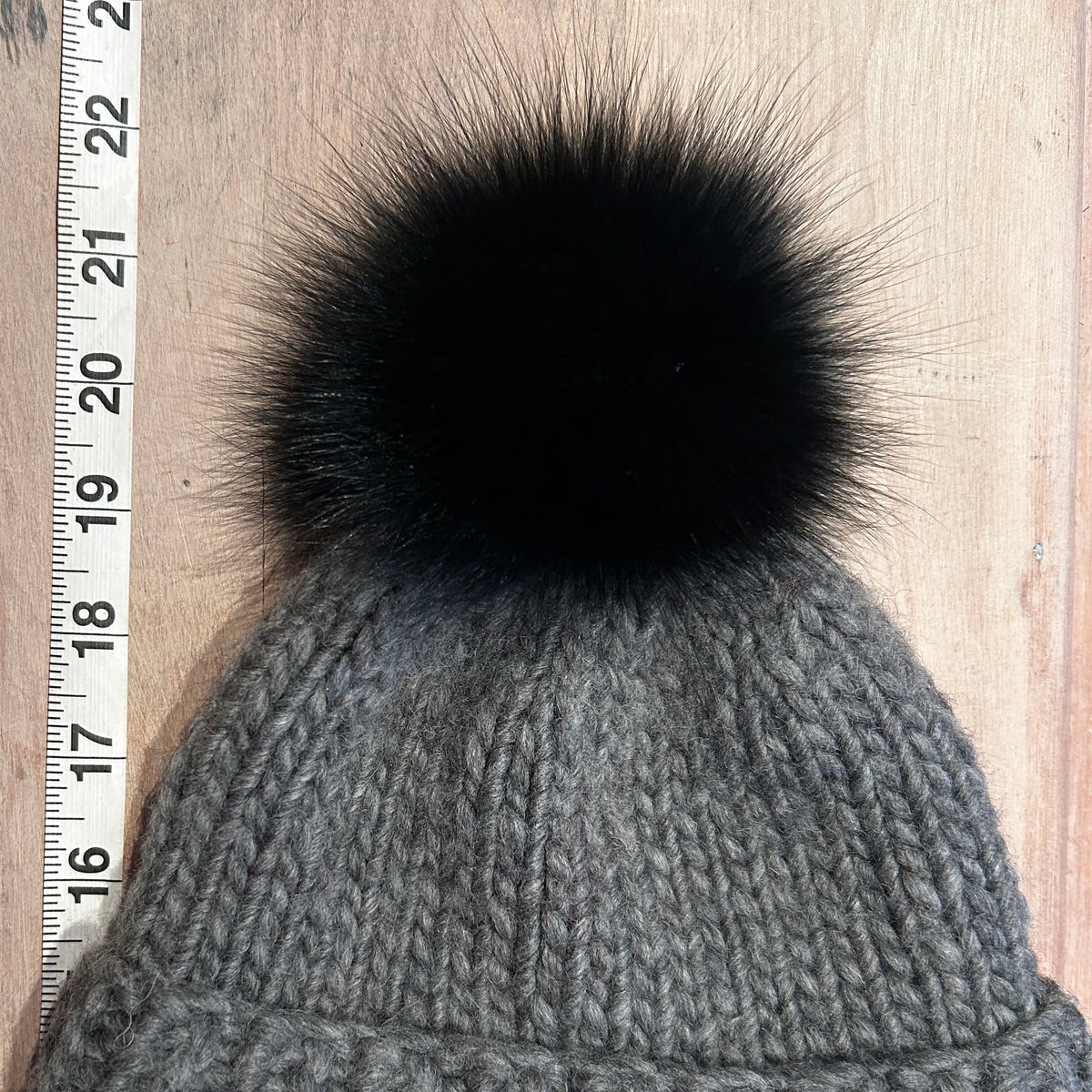 A small gray ribbed knit beanie with a small black faux-fur pom-pom, shown flat on a light wood surface next to a measuring tape.