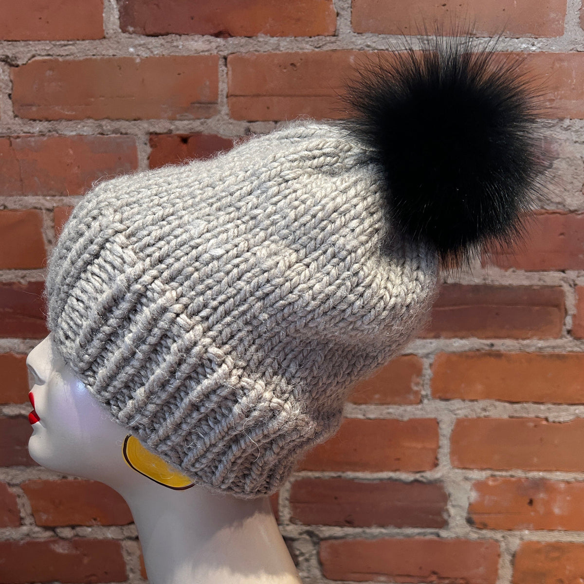 A light gray ribbed knit beanie with a large black fox-fur pom-pom, displayed on a white mannequin head against a red-brown brick wall background.