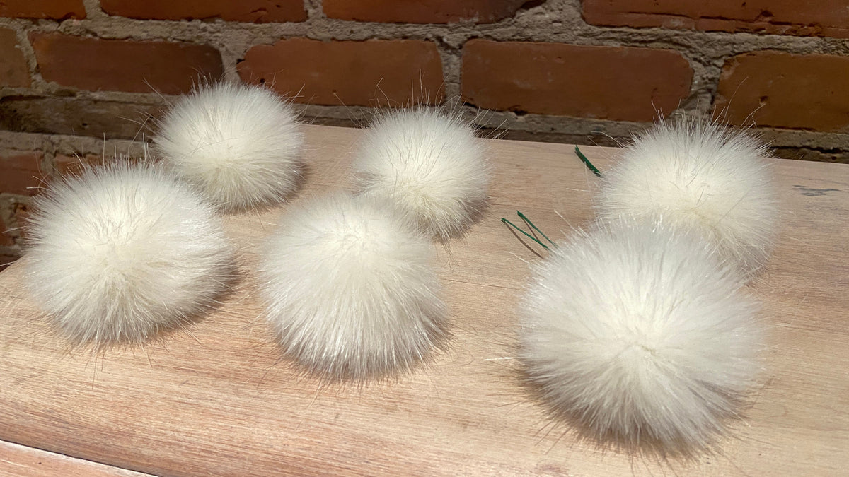 Six fluffy white faux fur pom-poms arranged on a light wood surface with a brick wall background.