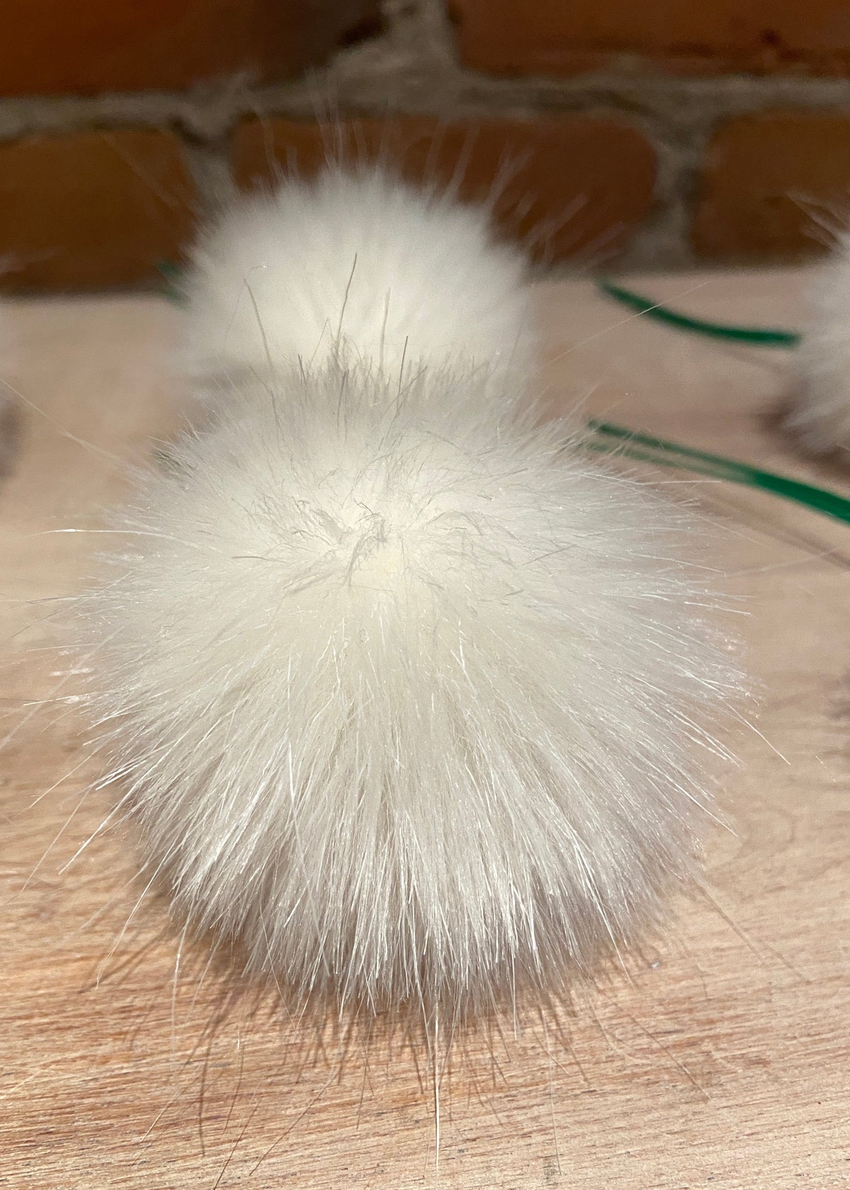 A close-up of a fluffy white faux fur pom-pom accessory on a light wood surface with a brick wall background.