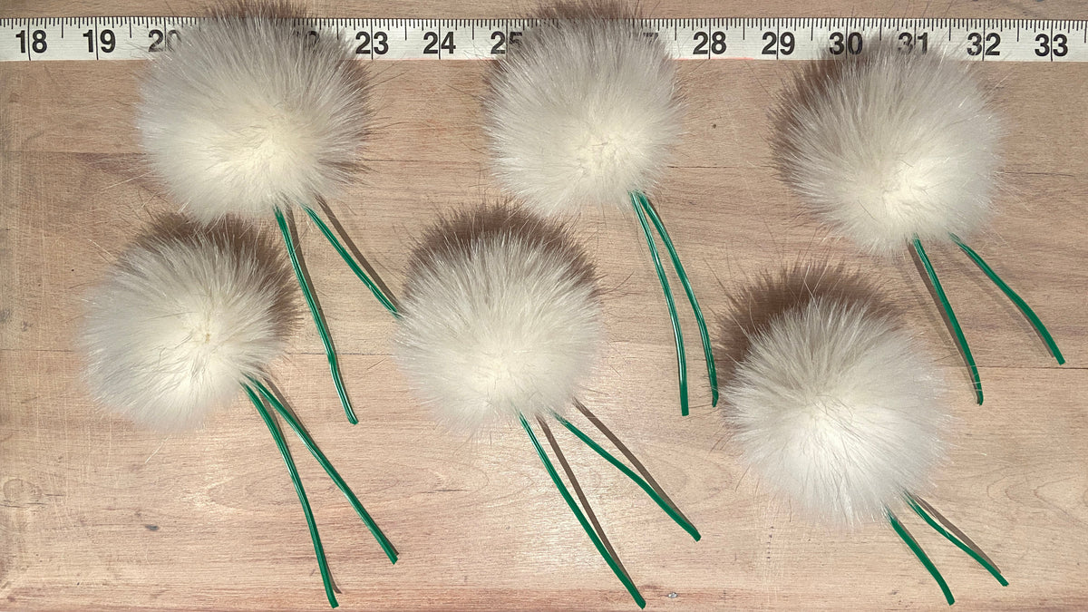 Five white faux fur pom-poms with green wire stems arranged on a light wood surface next to a metal ruler for scale.