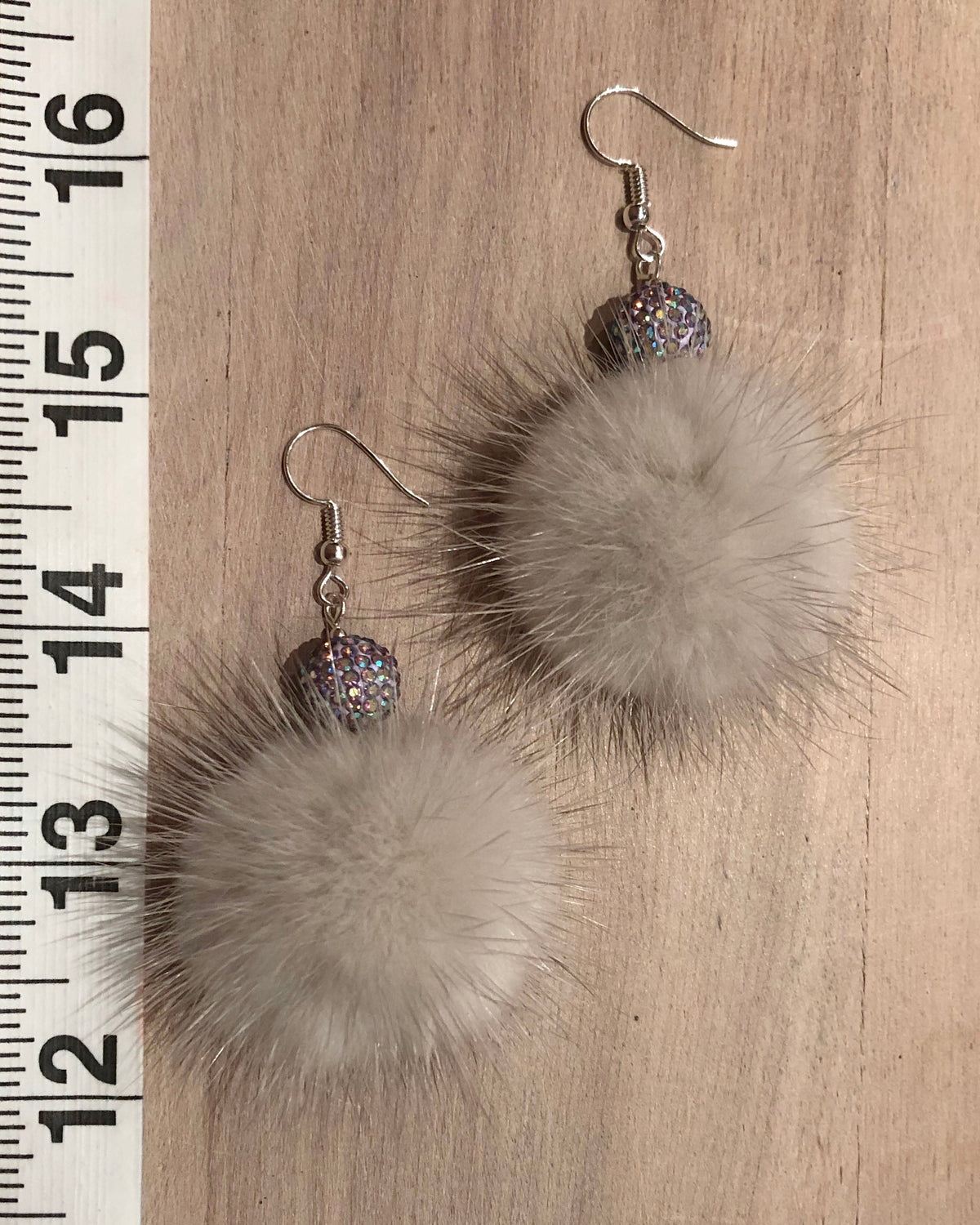 A pair of handmade dangle earrings featuring large, fluffy light grey mink fur pom-poms, a small round iridescent glitter bead, and silver fishhook ear wires, displayed on a wooden surface next to a measuring ruler.