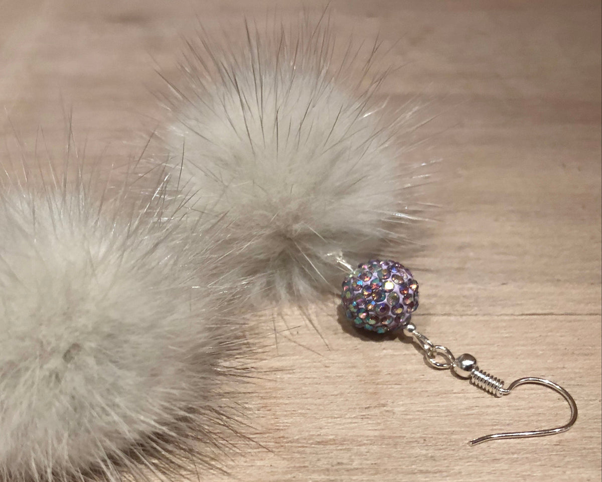 A pair of fluffy pom-pom earrings made from light grey fur, with a small iridescent rhinestone bead and silver fishhook wires, displayed on a light wood surface.