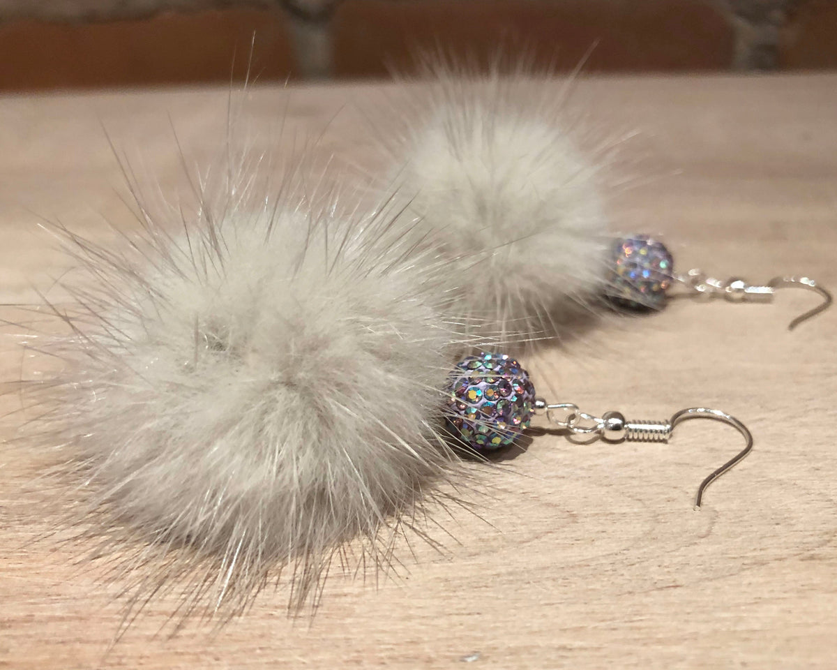 A pair of handmade dangle earrings featuring large, fluffy light gray mink fur pom-poms and a small iridescent glittery bead, hanging from silver fishhook wires on a light wood surface.