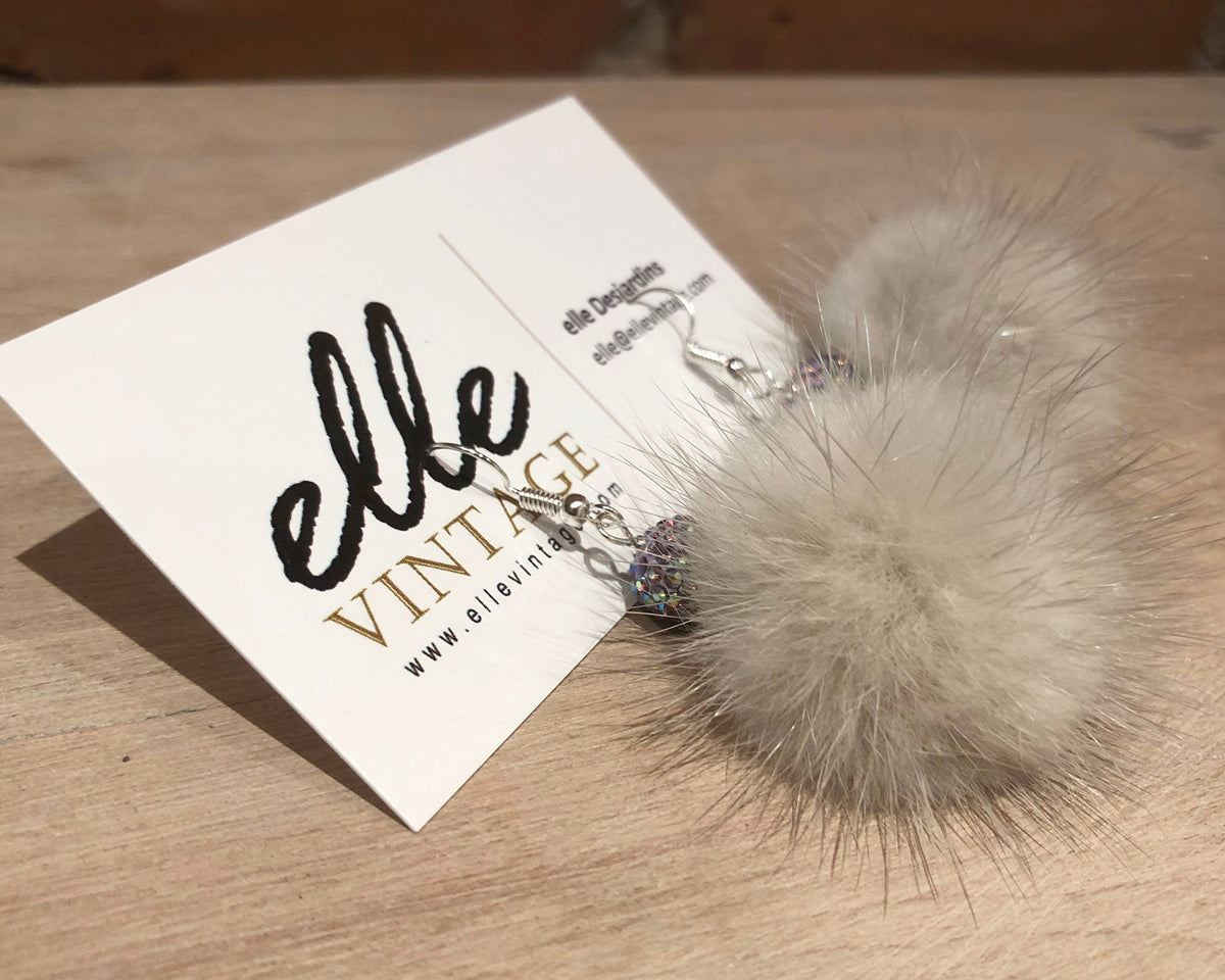 A pair of dangle earrings with fluffy cream mink fur balls and small iridescent rhinestone charms on silver hooks, displayed on a white card labeled 'elle VINTAGE' on a wooden surface.