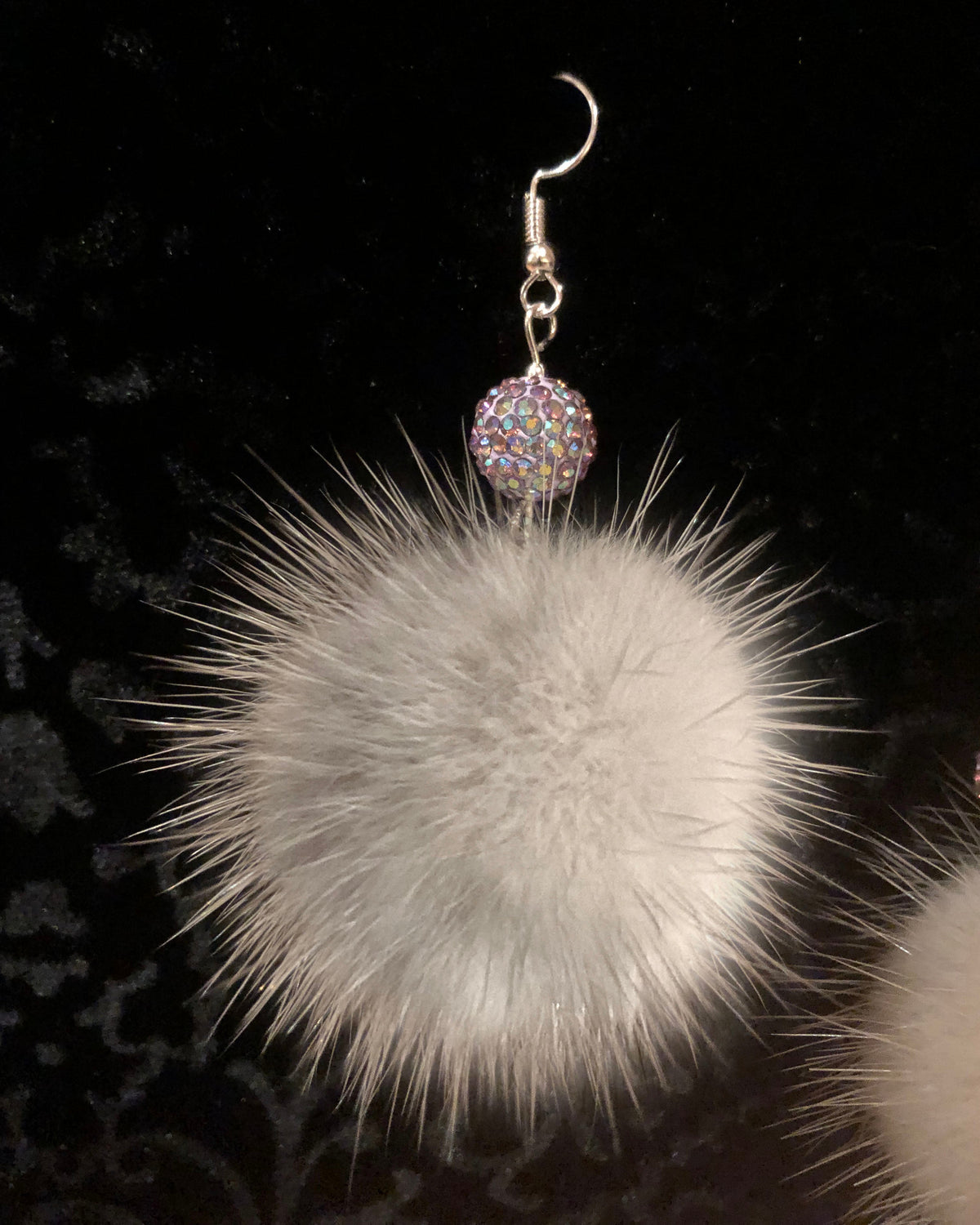 A close-up of a fluffy white mink fur pom-pom earring with a silver-tone hook and a sparkling iridescent rhinestone bead at the top, set against a black background.
