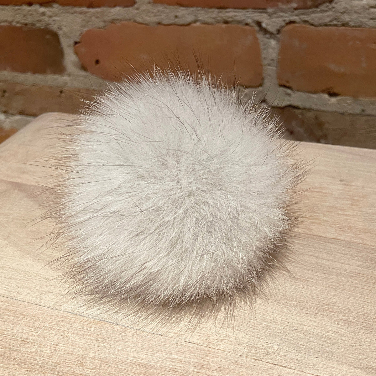 A fluffy white fox fur pom-pom ball resting on a light wood surface with a brick wall background.
