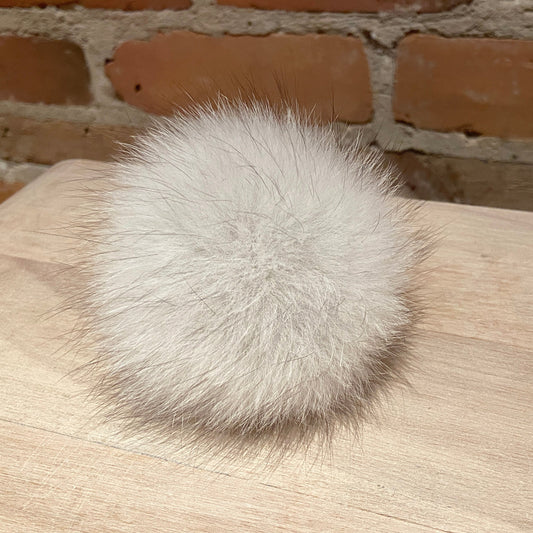 A fluffy white fox fur pom-pom ball resting on a light wood surface with a brick wall background.
