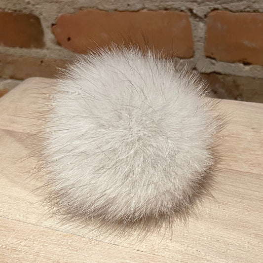 A fluffy white fox fur pom-pom ball resting on a light wood surface with a brick wall background.